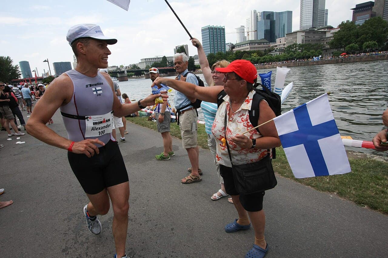 Alexander Stubb running at Ironman Frankfurt in 2009, in a Finland race suit with the number 100, as a supporter waving a Finnish flag hands him a drink.