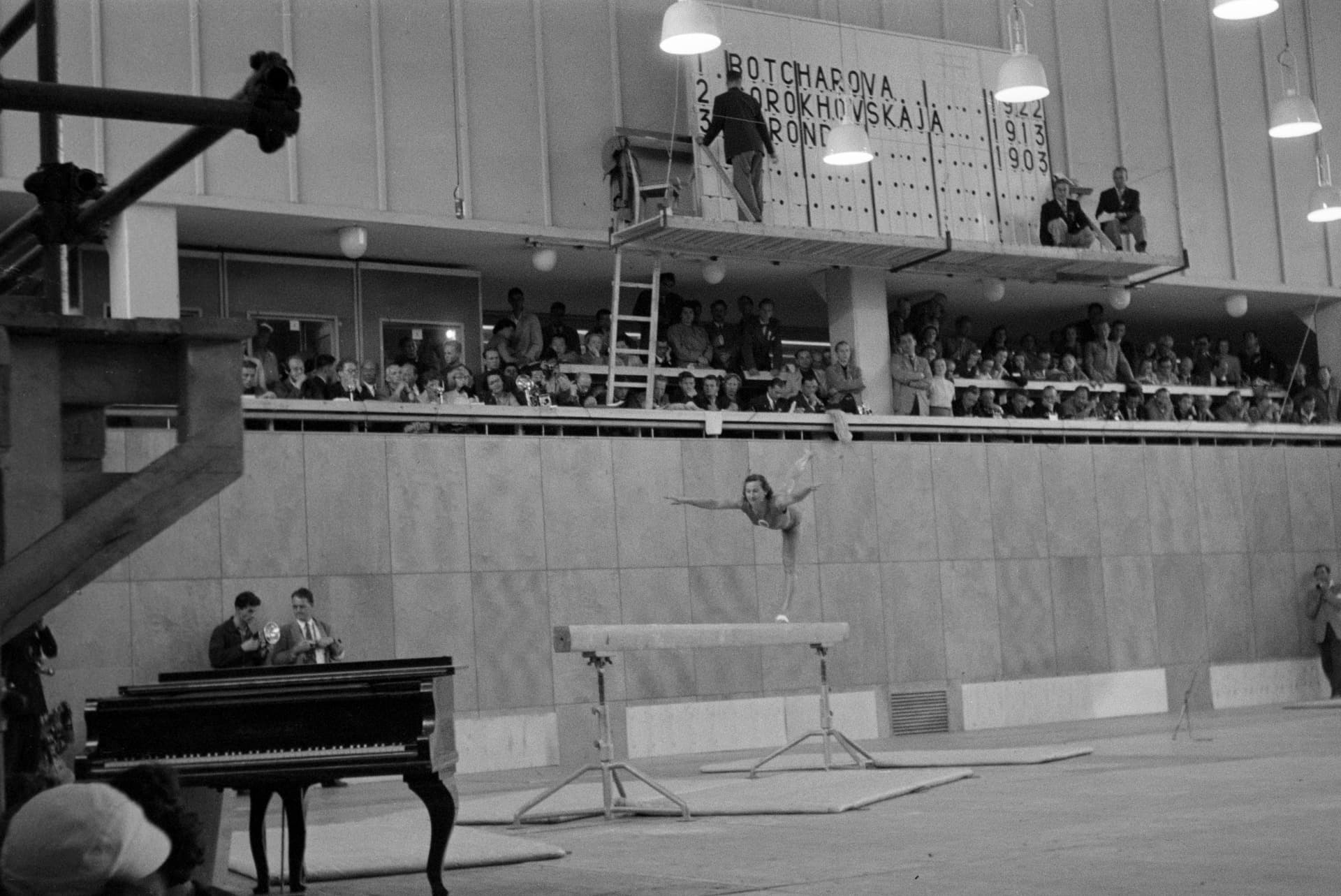 A black and white photograph of a gymnast on the balance beam at the 1952 Olympic Games in Helsinki. She stands on one leg on the narrow beam, tipped forward with her back leg lifted high behind her and her arms spread wide, while the crowd and the judges watch.