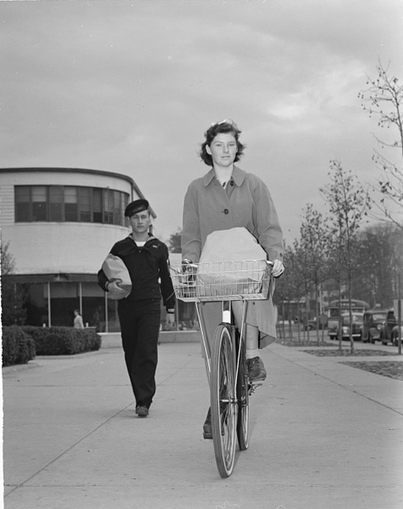 A young woman rides a bicycle towards the camera with a bag of groceries in its front basket, while a sailor walks behind her carrying a paper bag of shopping under his arm. Greenbelt, Maryland, 1942.