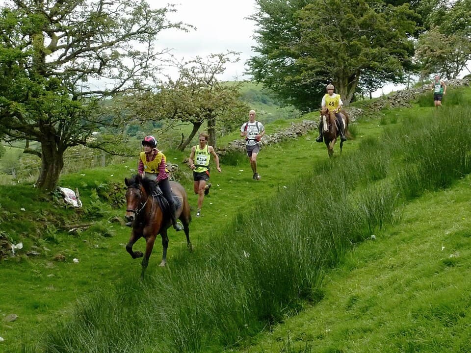 Runners and riders on horseback on a green track in the Welsh hills during the Man versus Horse race.