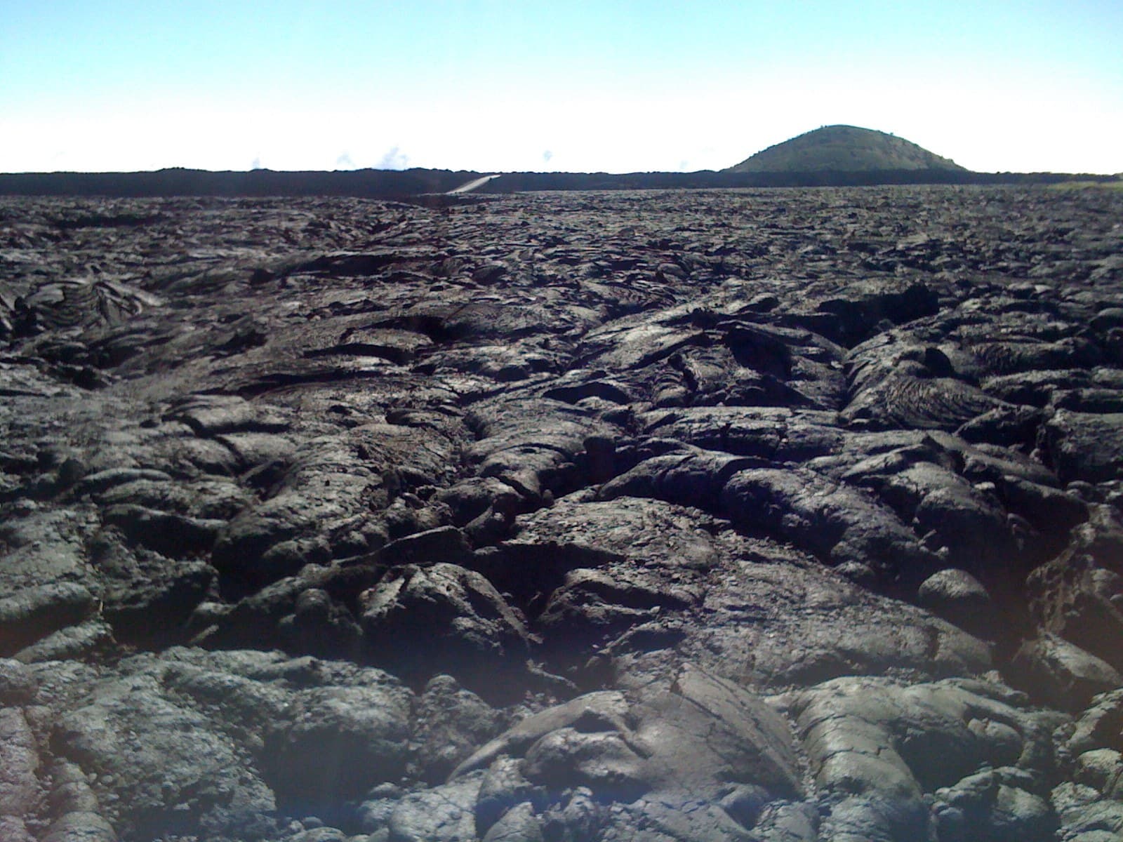 A field of black lava from Mauna Loa running to the horizon, a cinder cone behind.