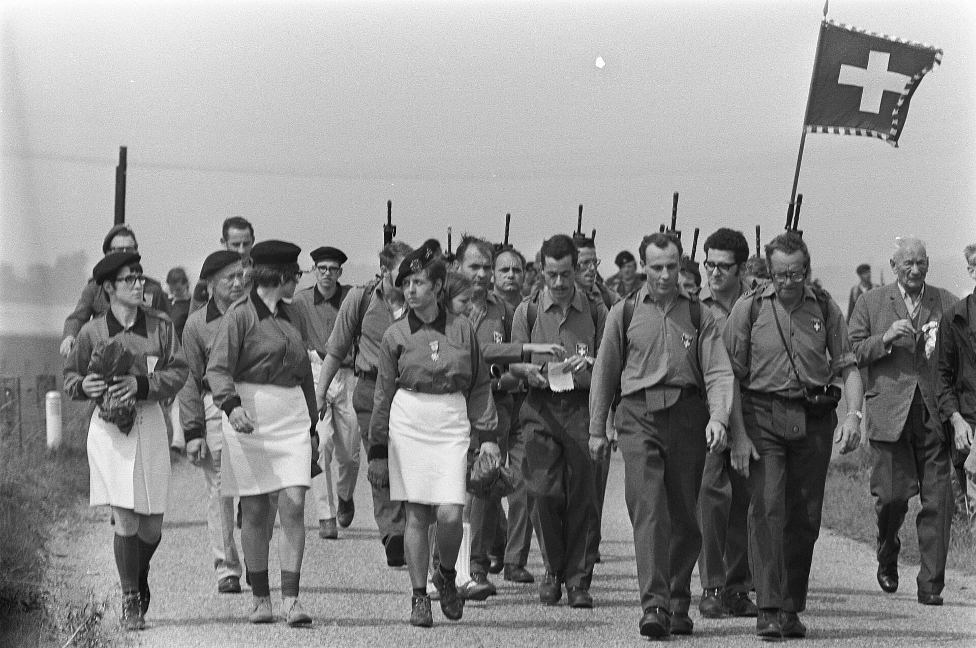 Walkers in the 1970 Nijmegen Four Days March behind a Swiss flag, some soldiers with rifles.