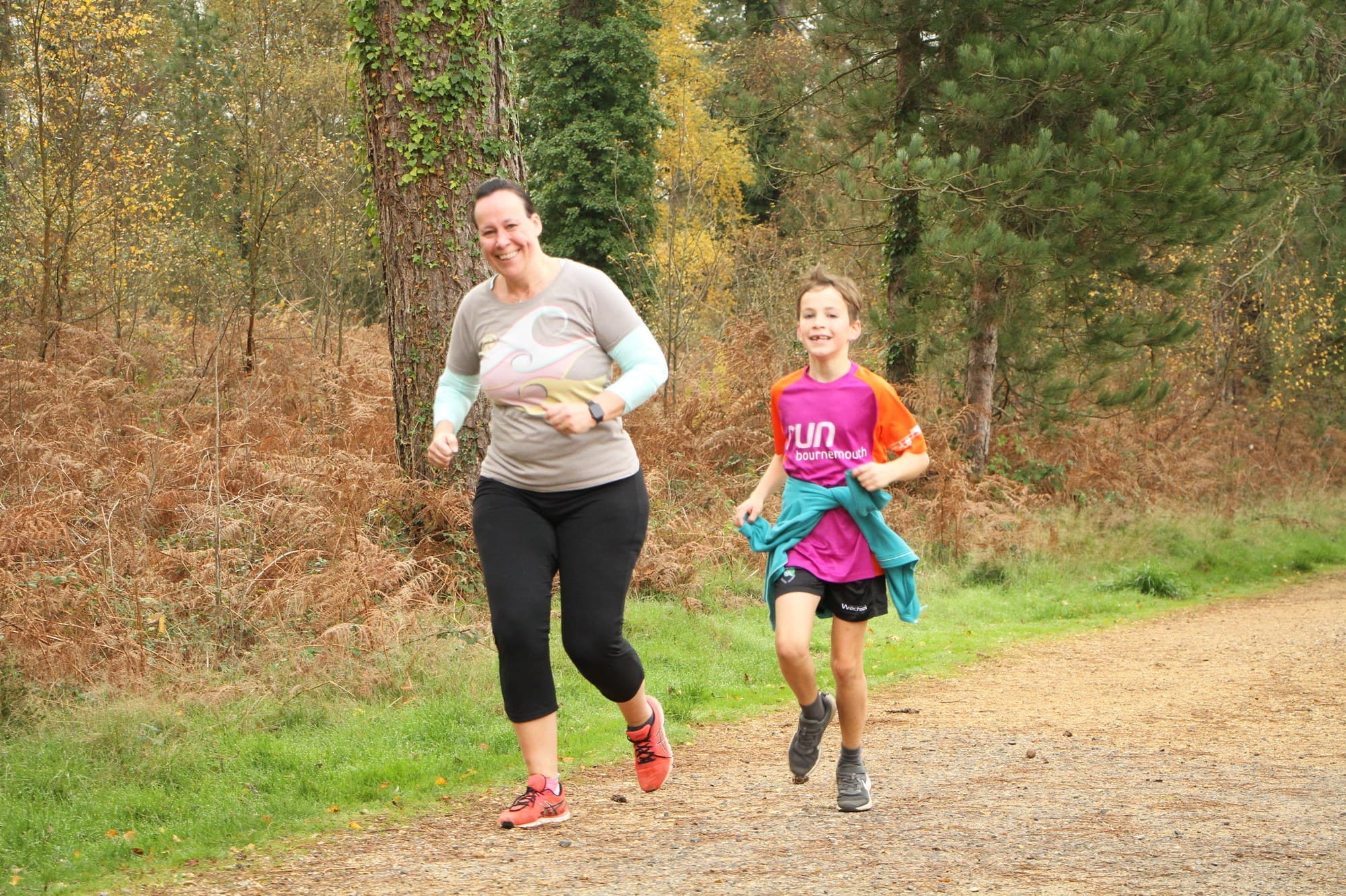 A woman and a girl running together on a woodland path at a parkrun.