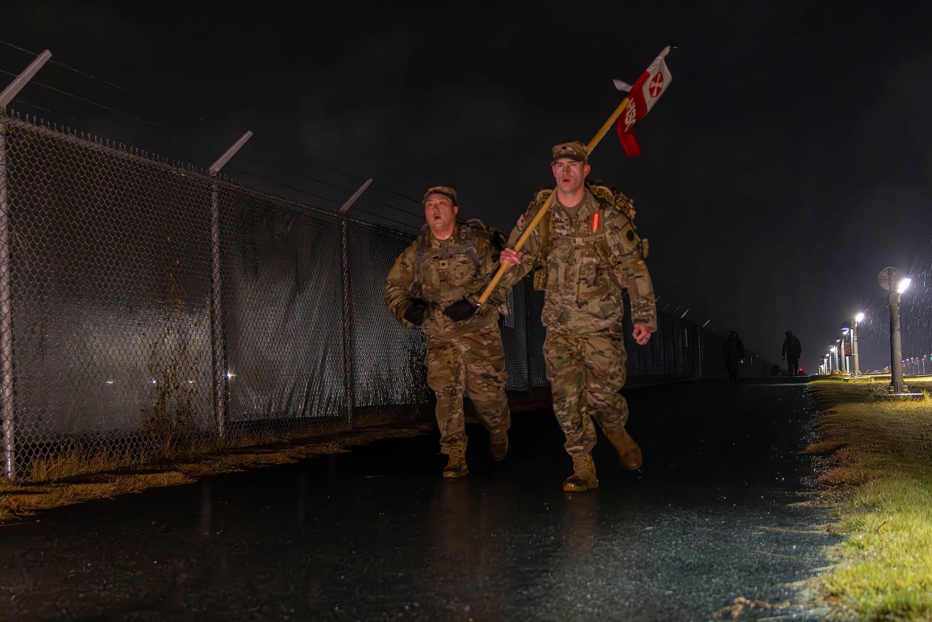 Two soldiers with rucksacks and a guidon on a 12-mile foot march before dawn.