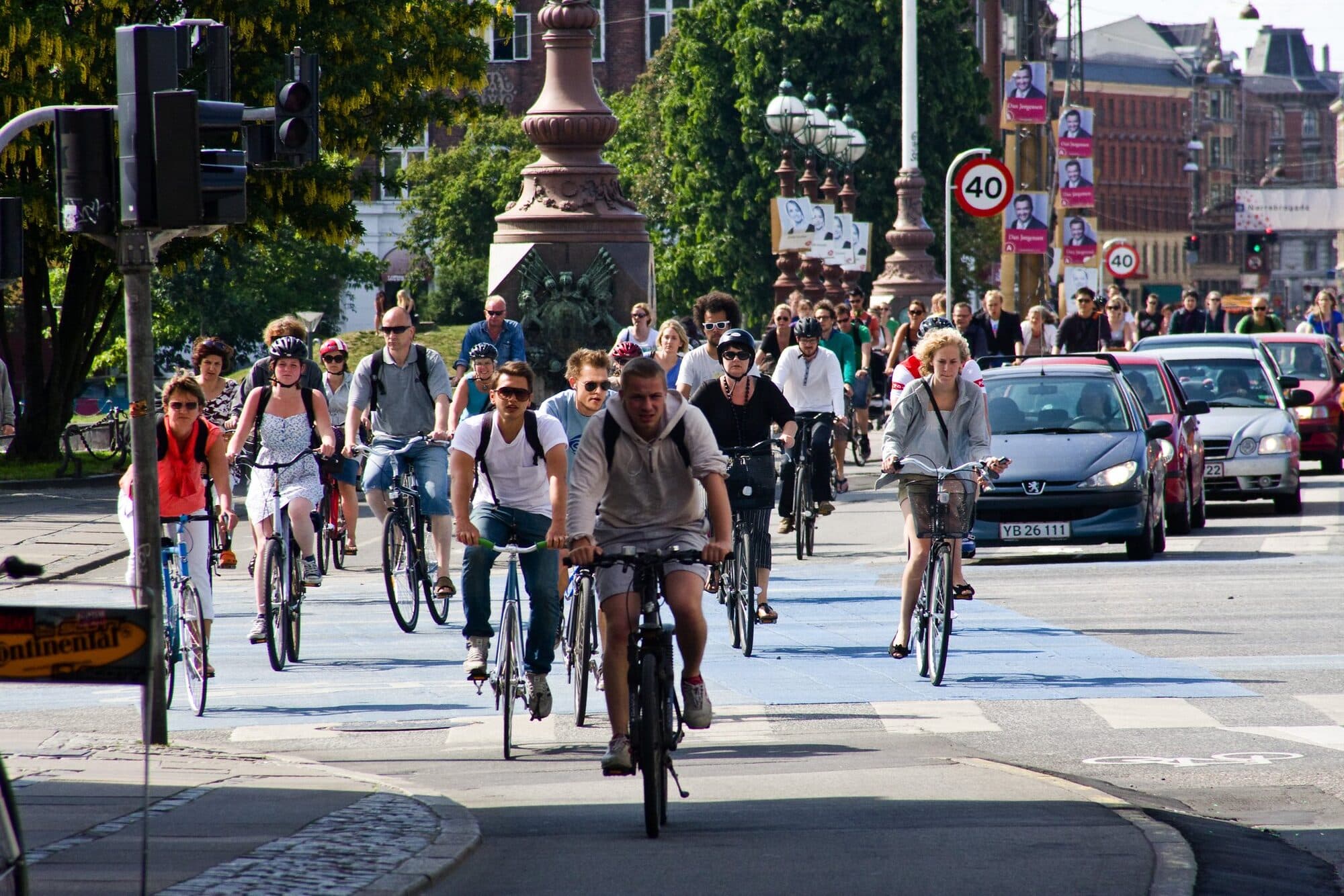 A crowd of people in everyday clothes cycle through a Copenhagen junction on a sunny afternoon, with bags on their backs and in their baskets, beside cars waiting at the lights.