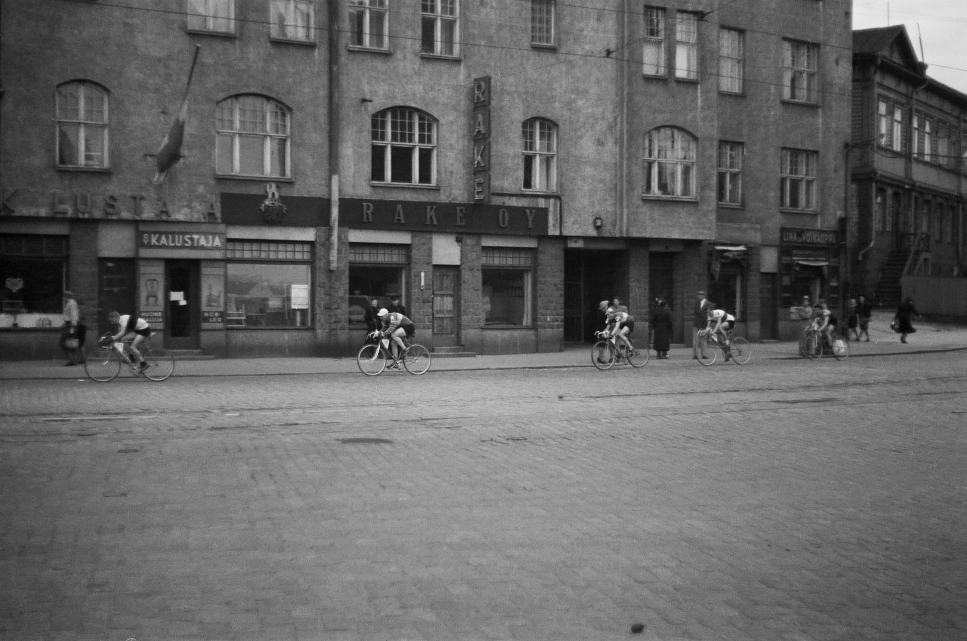 A black and white photograph from 1954 of racing cyclists strung out along a city street, riding fast over a wide expanse of rough cobblestones. Each rider sits low and steady over the bars while the stones jolt the wheels beneath them.
