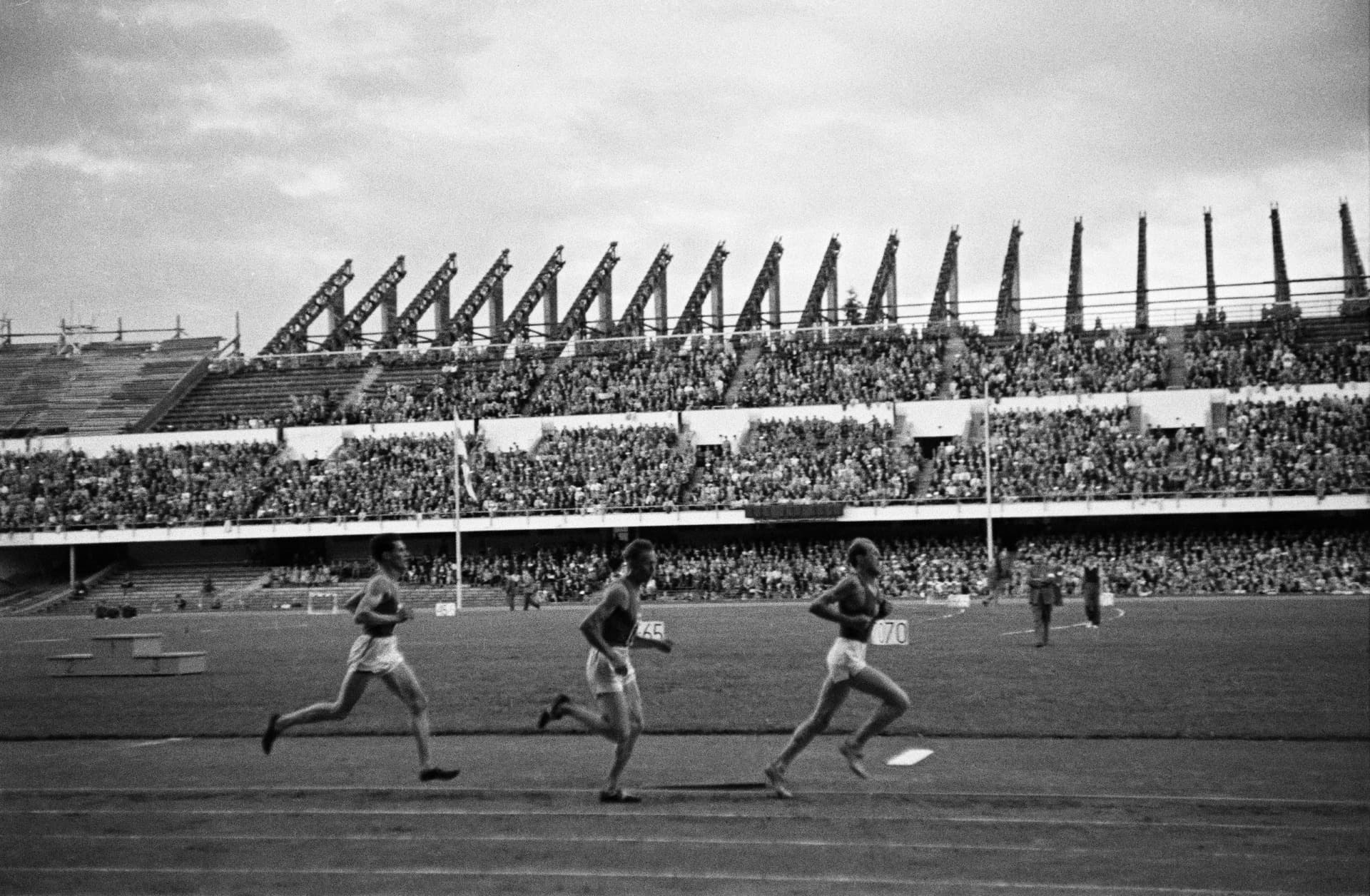 A black and white photograph from 1949 of three distance runners in profile on the track of the Helsinki Olympic Stadium, in front of a packed stand. With each stride the back leg swings out long behind the runner, the hip opening as the foot pushes away.