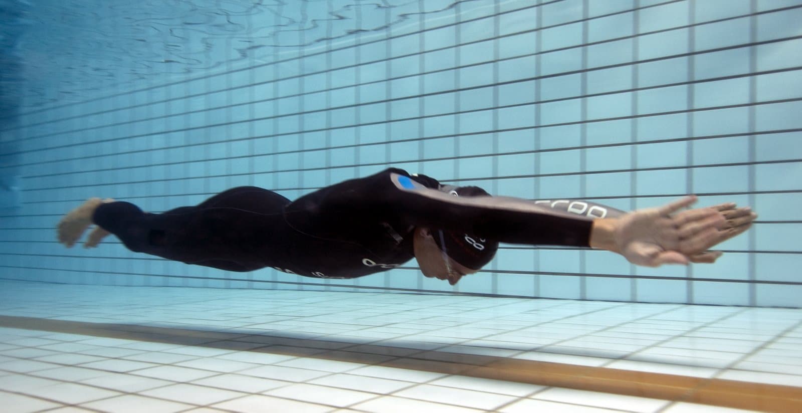 A freediver in a black wetsuit glides underwater just above the floor of a pool, seen from the side. His arms reach straight out beyond his head, his head is tucked between them, and his body is one straight line from fingertips to feet.