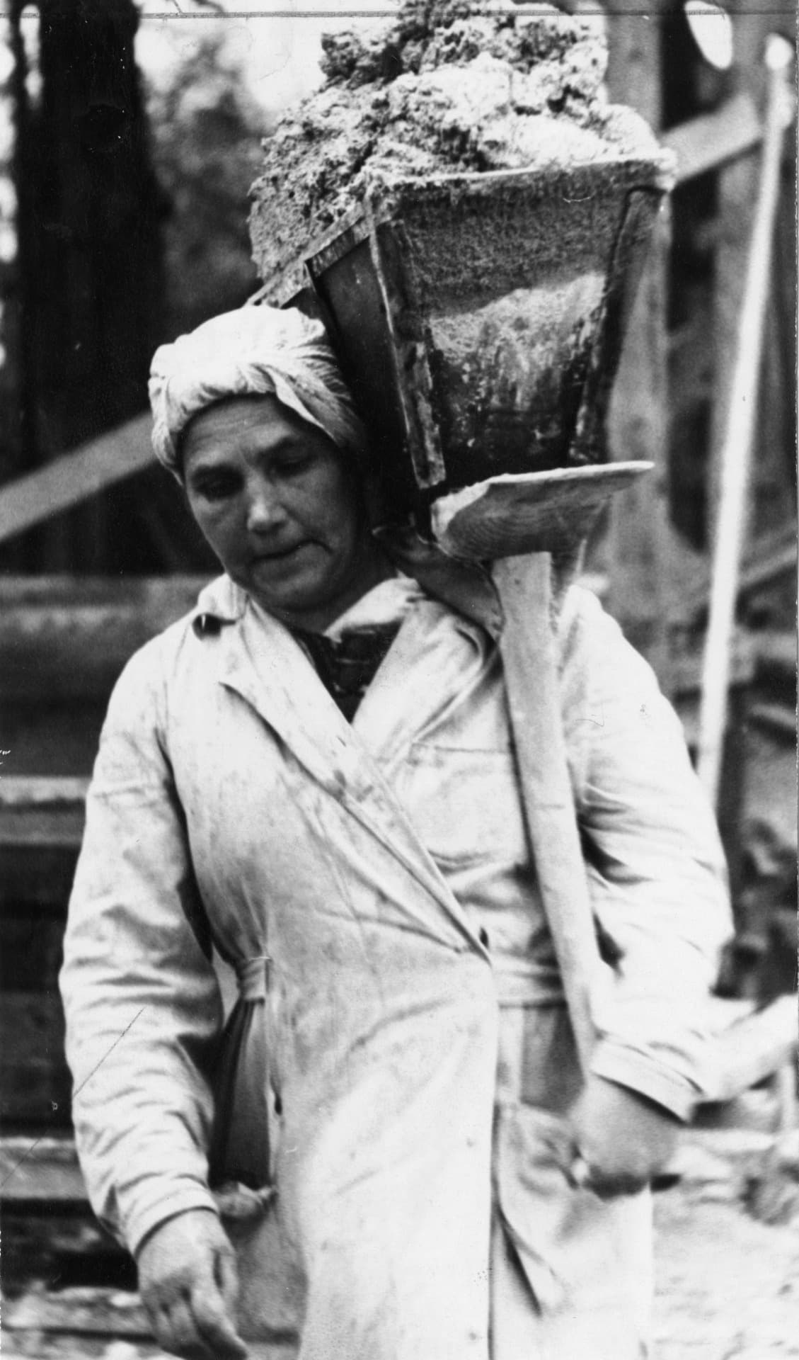 A black and white photograph from 1949 of a woman on a building site carrying a heavy hod of mortar on one shoulder. Under the load her back stays long and her chest stays up, one hand steadying the handle in front of her as she walks.