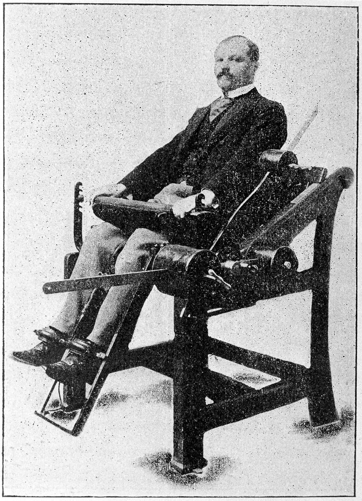 An old photograph of a man in a suit sitting in a heavy iron exercise chair. His back is supported and his shins are strapped under a padded bar on a lever, so only his knees can move.