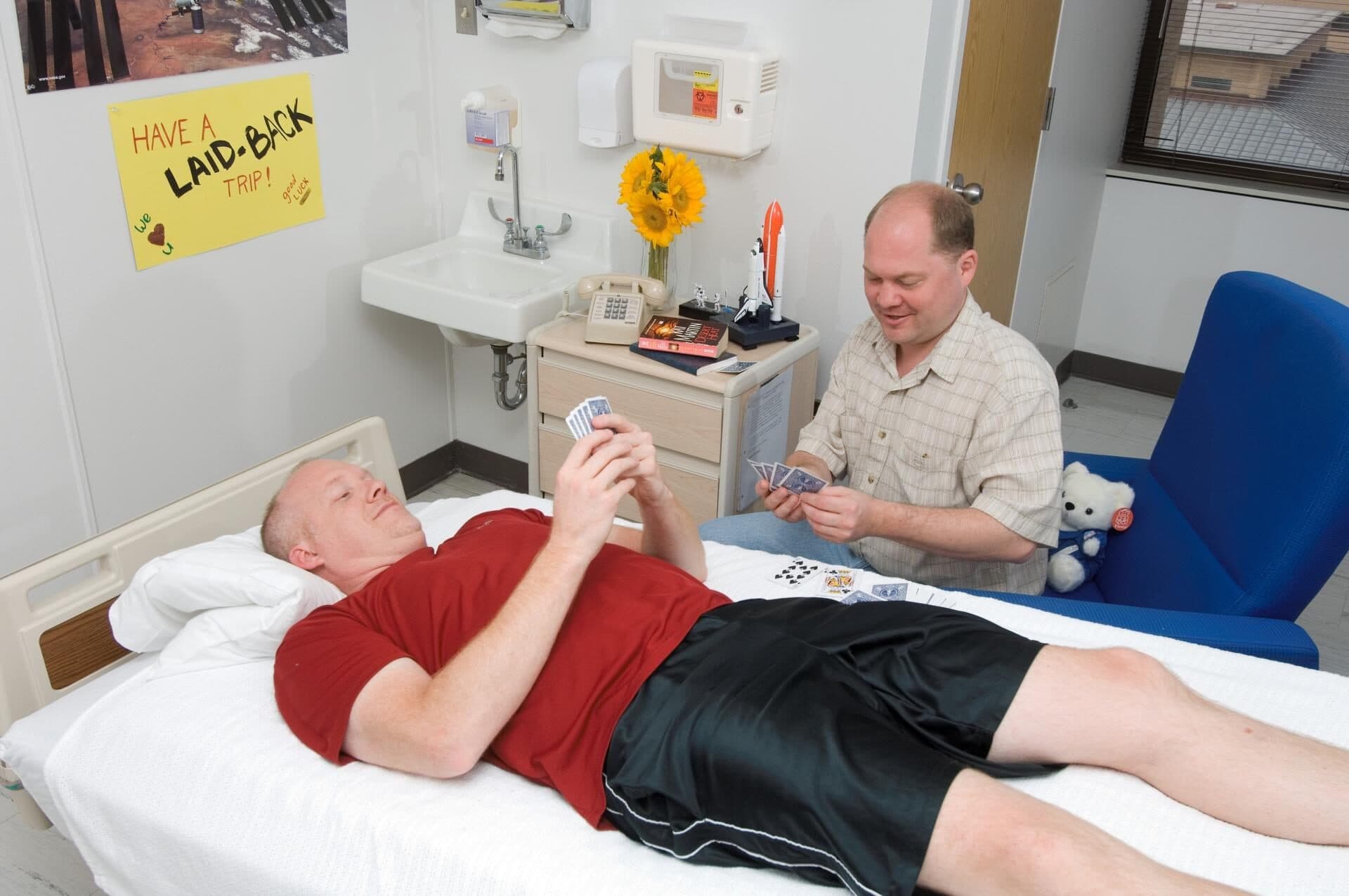 A healthy young man in a red T-shirt lies flat on a hospital bed, playing cards with a visitor. He is a volunteer in a NASA bed rest study in Galveston, Texas, and a sign on the wall reads: Have a laid-back trip.