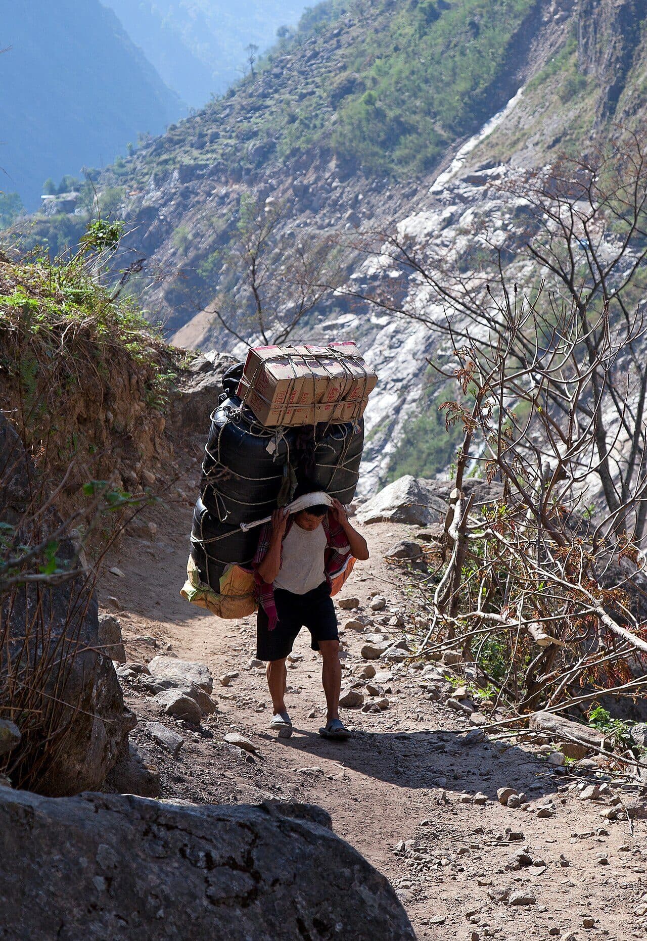 A porter climbs a rocky mountain trail in Nepal in sandals, carrying a load of bags and boxes taller than he is. A strap across the top of his head holds it.