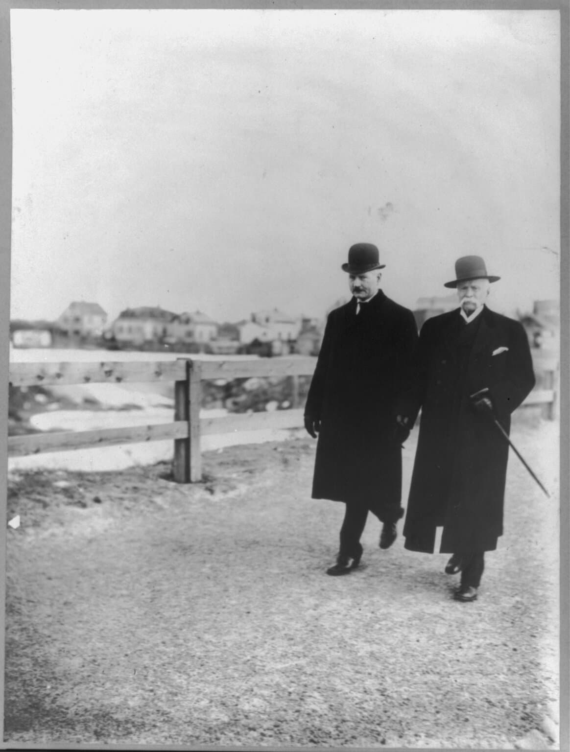 Two men in long dark coats and hats walk briskly along a gravel road beside a wooden fence. The one on the right, white-moustached and carrying a cane, is the walker Edward Payson Weston, then in his late sixties.