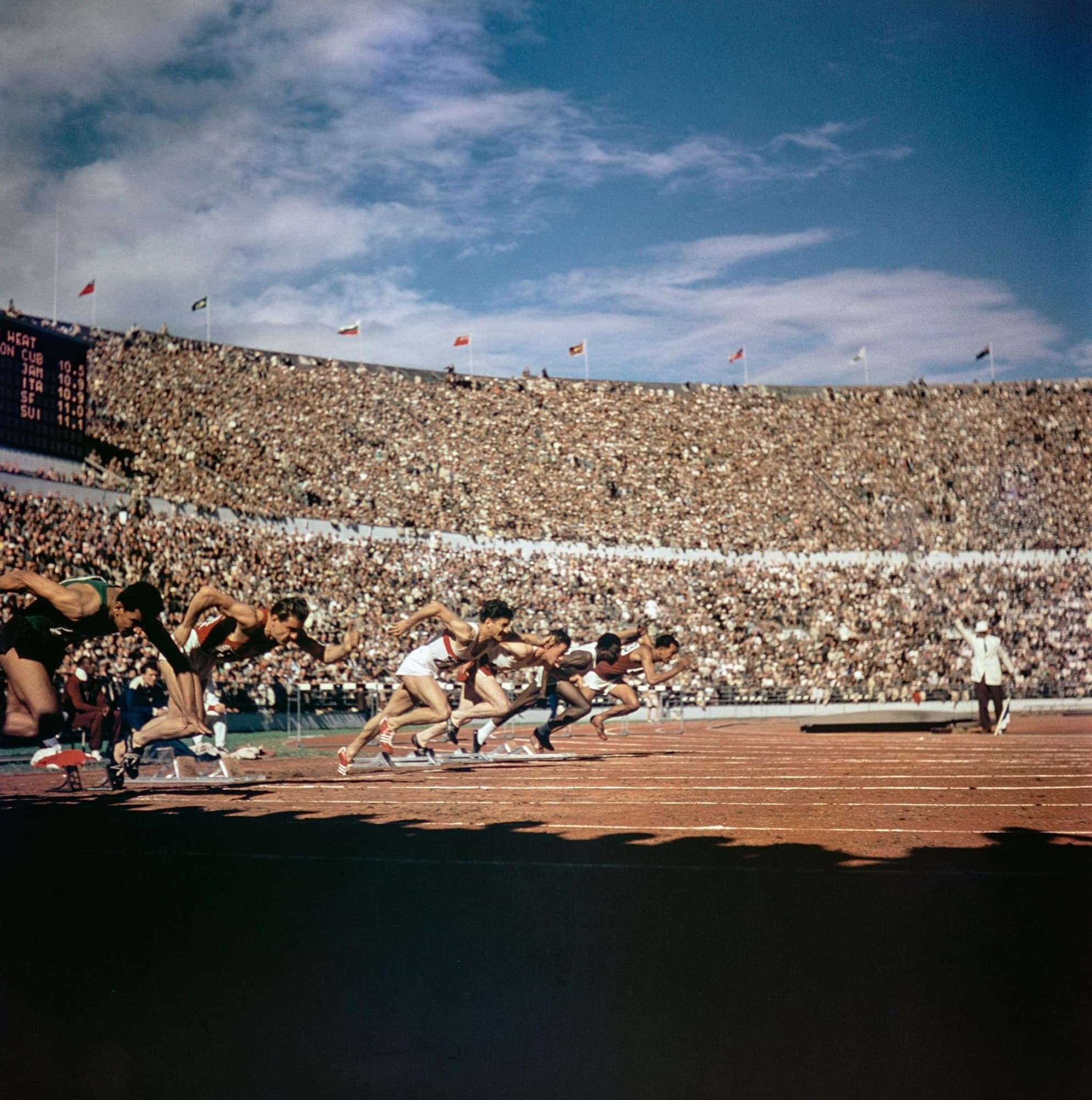 A colour photograph of a heat of the 100 metres at the 1952 Olympic Games in Helsinki. A line of sprinters drives out of the starting blocks in front of a packed stand, bodies low and leaning far forward, arms pumping and back legs pushing hard off the blocks.