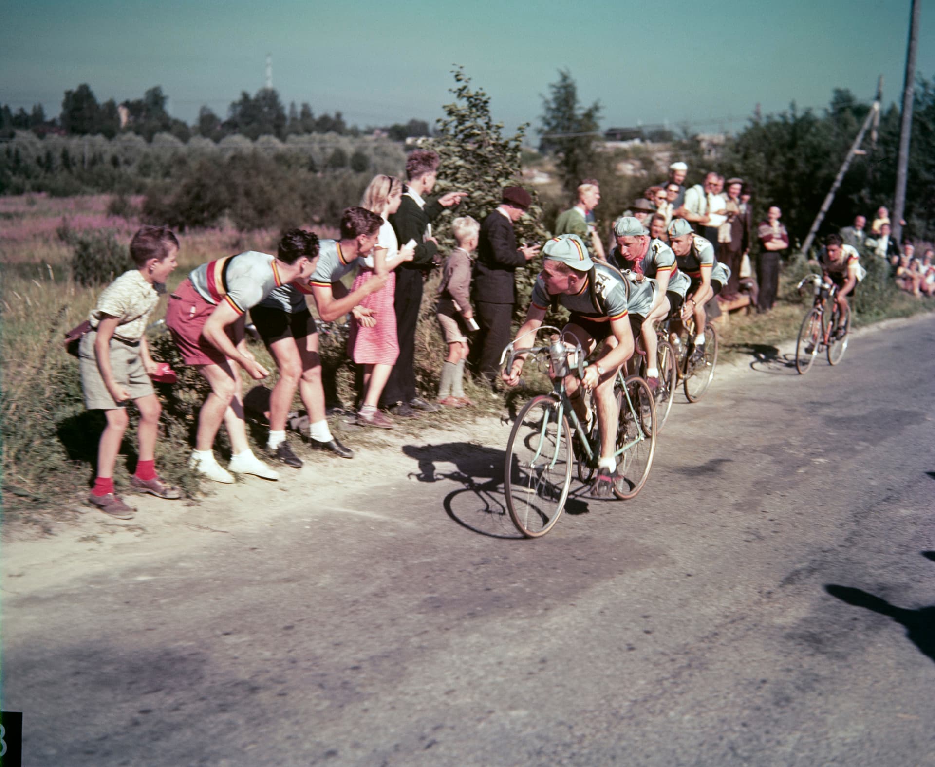 A colour photograph of the 1952 Olympic road race in Helsinki. Riders on a dusty road ride in a close line, each on the wheel of the one ahead, while spectators and boys in shorts lean in from the verge to watch them pass.