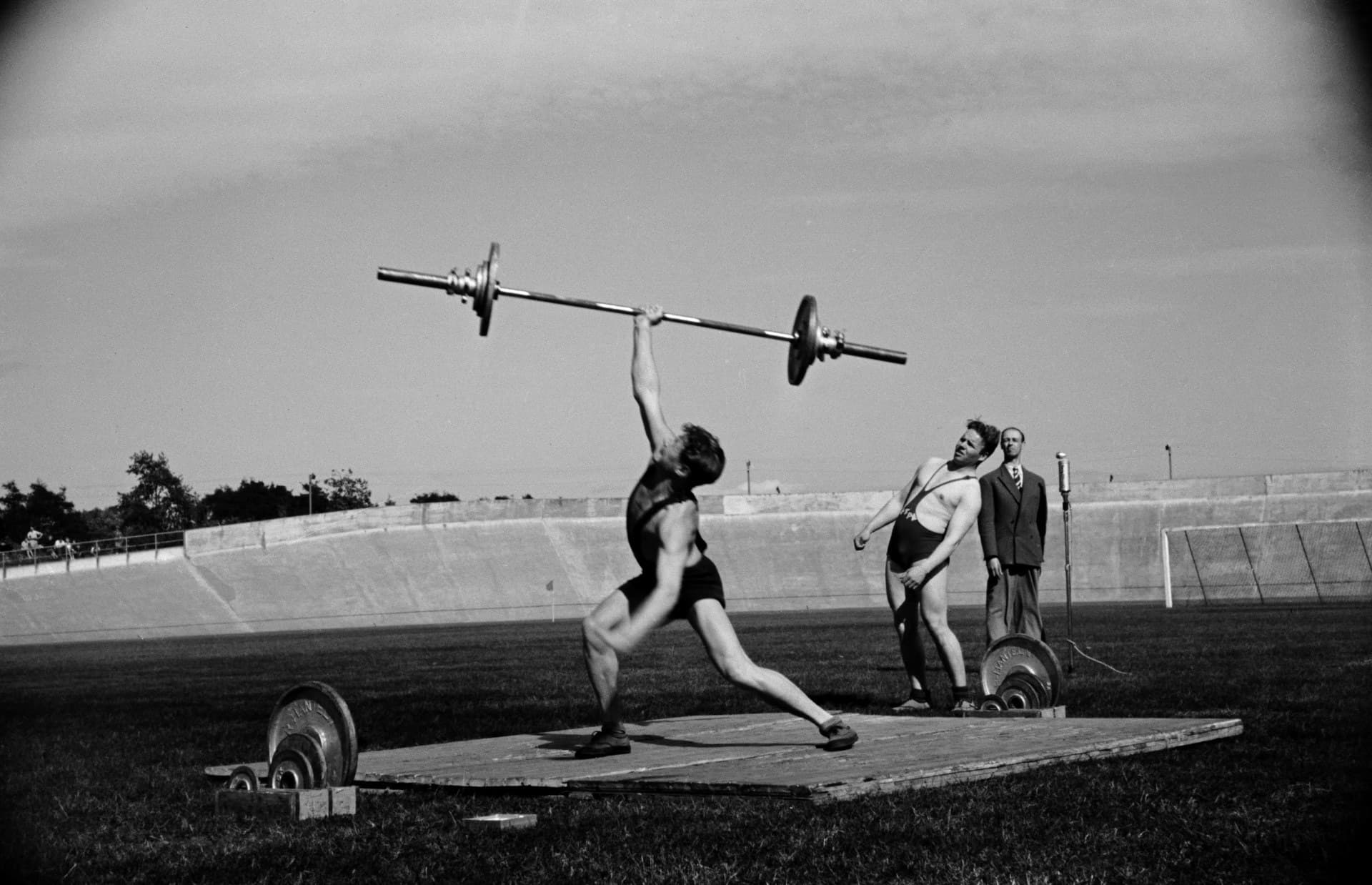 A black and white photograph from 1948 of a weightlifter on a wooden platform in an open-air velodrome, catching a barbell overhead on one straight arm. He has dropped into a deep split under the bar, front knee bent and back leg stretched long, while another lifter and an official watch.