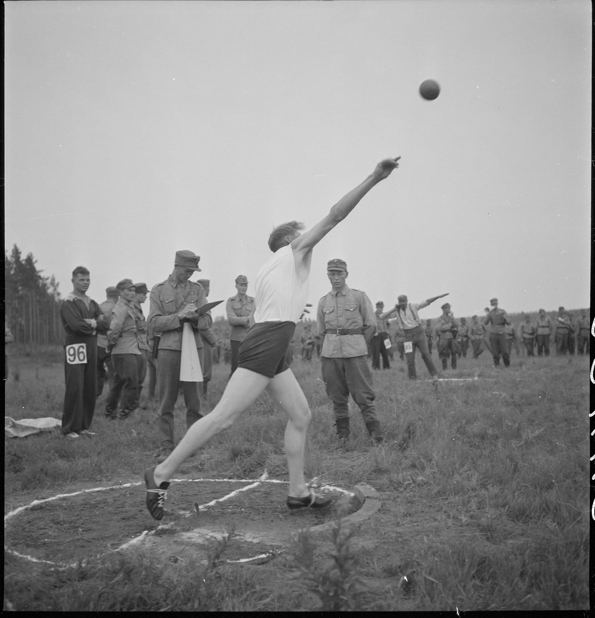 A black and white photograph from 1942 of a young man in a white vest and dark shorts putting the shot at an outdoor sports day. His back foot drives off the edge of the circle, his hips and chest have turned towards the throw, and his arm is stretched out full length after the shot, which flies away above the watching crowd.
