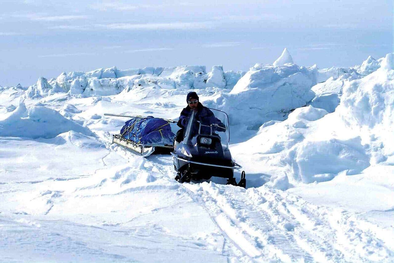A snowmobile pulls a loaded wooden sled across rough sea ice on Baffin Island in the Canadian Arctic. One rider sits on the machine.