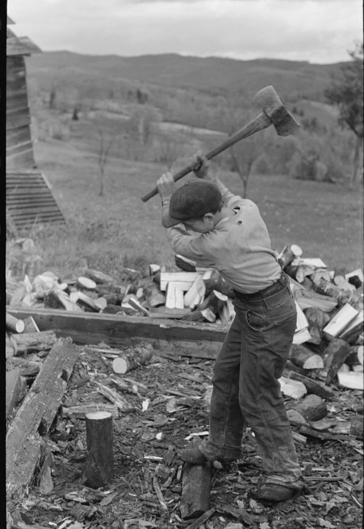 A boy on a Vermont farm swings an axe high over his head to split wood for the winter, feet planted wide among the chips, a pile of split logs behind him.