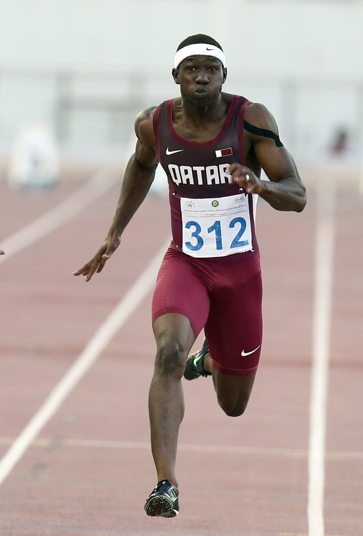A sprinter in a maroon Qatar vest drives down the track straight towards the camera, mid-stride in a 100 metres race. His arms pump hard, but his hands stay loose, one open and one lightly curled, never clenched.