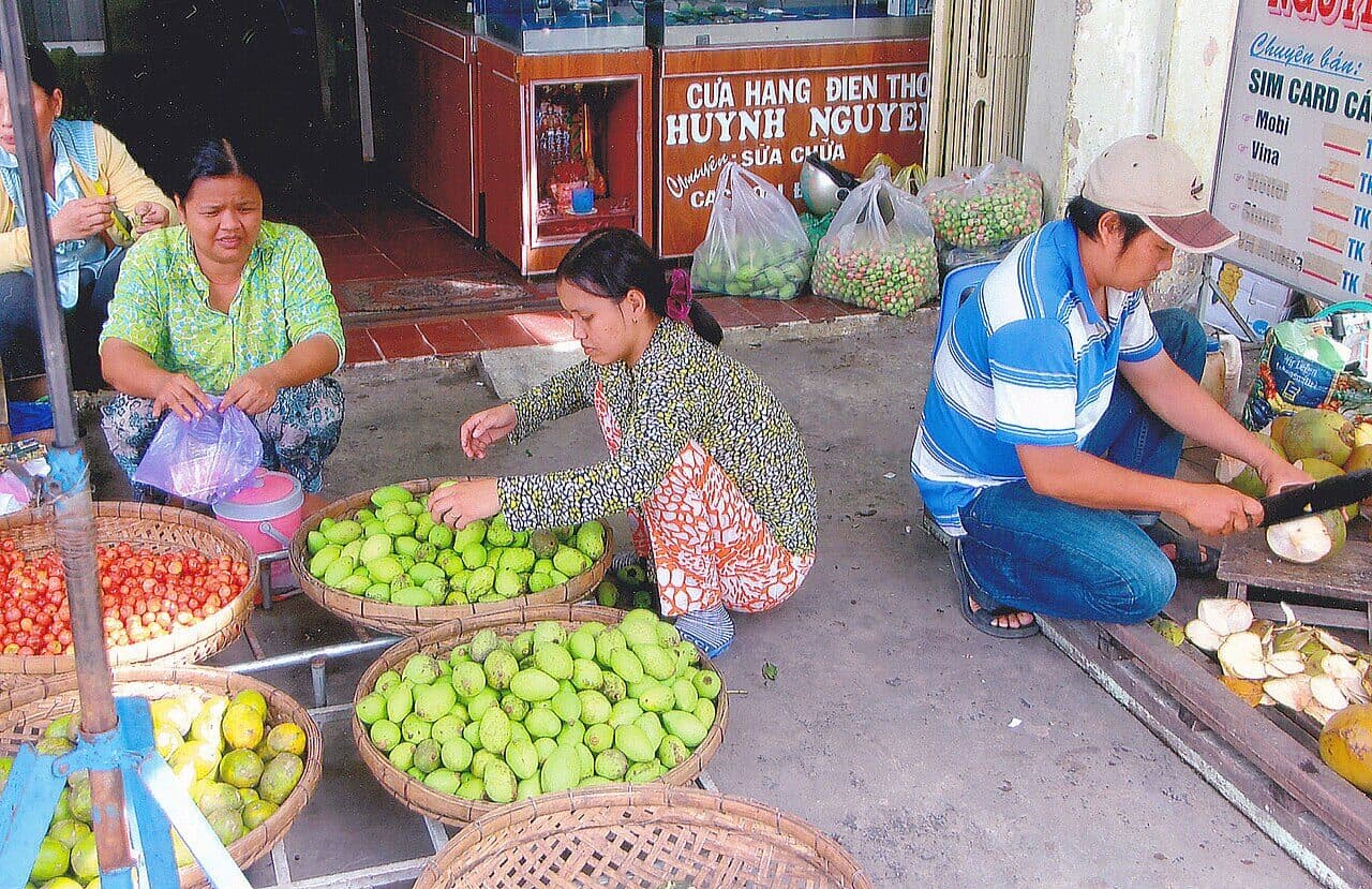 A street market in Vietnam. Two women and a man work at ground level in a deep squat, heels down, sorting fruit and cutting coconuts, with no stool or chair in sight.