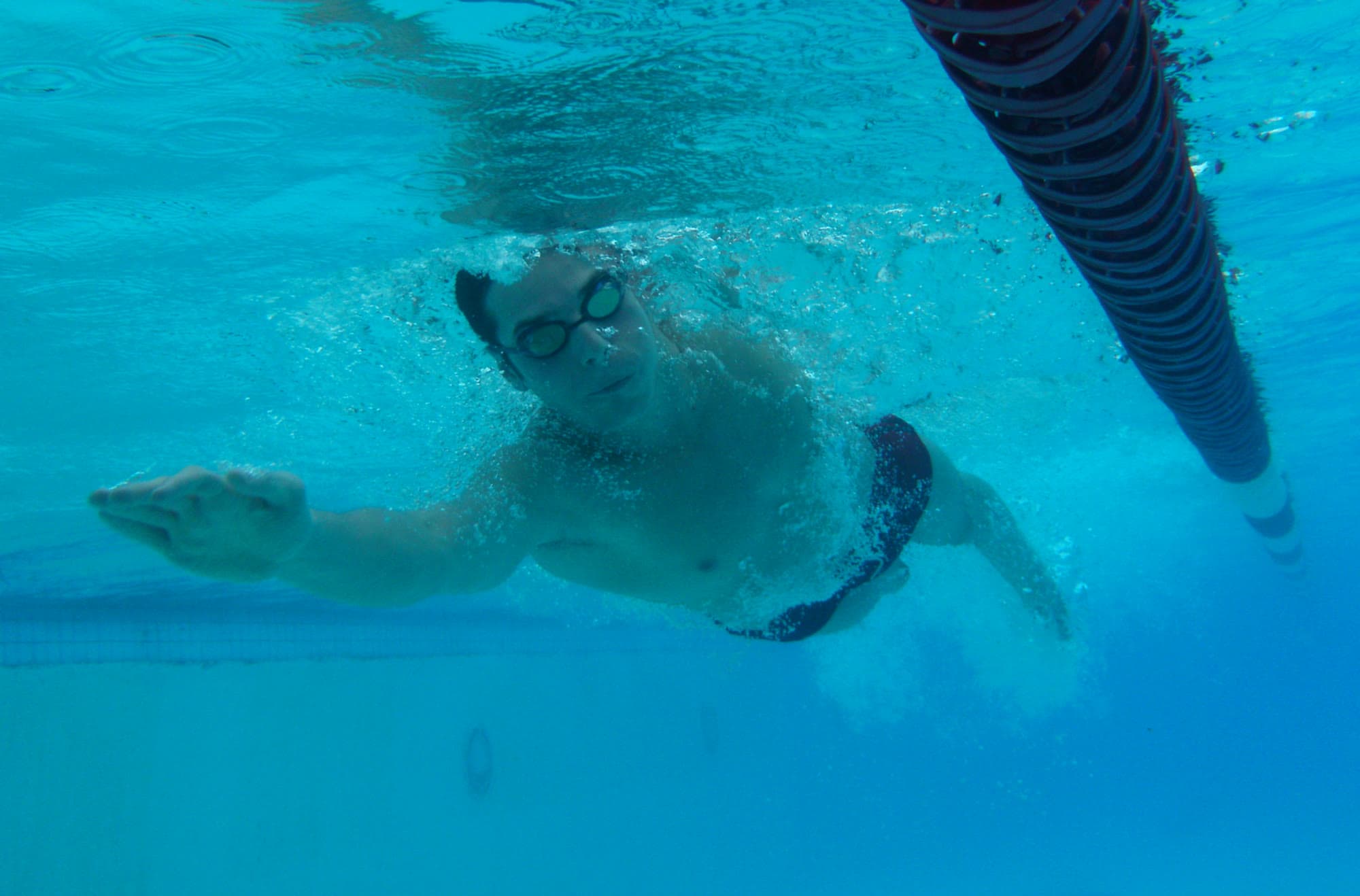 An underwater view of a swimmer doing front crawl in a pool beside a lane rope. One arm reaches straight forward from his shoulder, the hand flat and the fingers together, just starting to press back on the water.