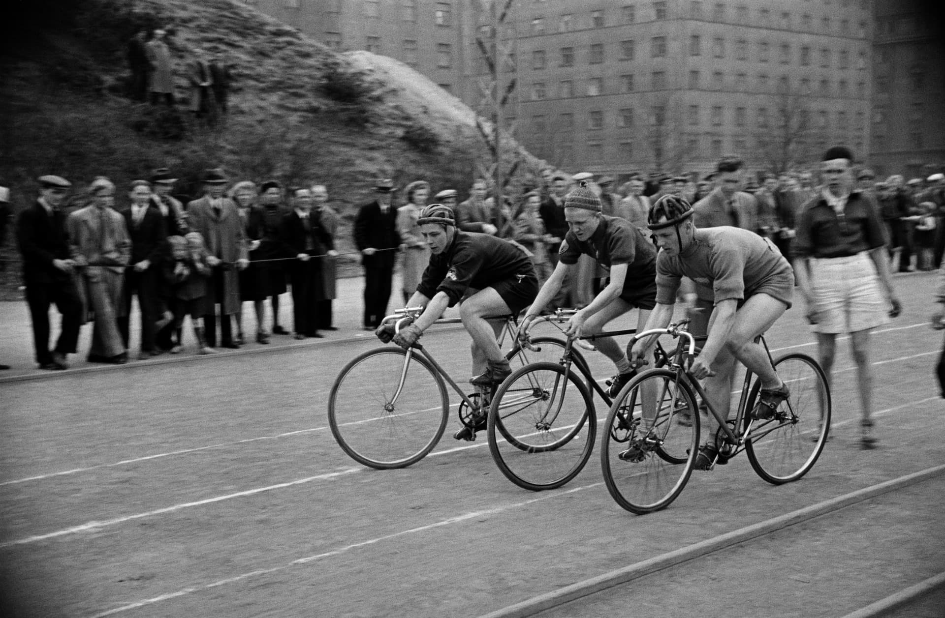 A black and white photograph from 1947 of three riders racing side by side at a sports ground in Helsinki, hands low on dropped handlebars. Each one leans far forward from the hips, back long and head up, while spectators line the track behind them.