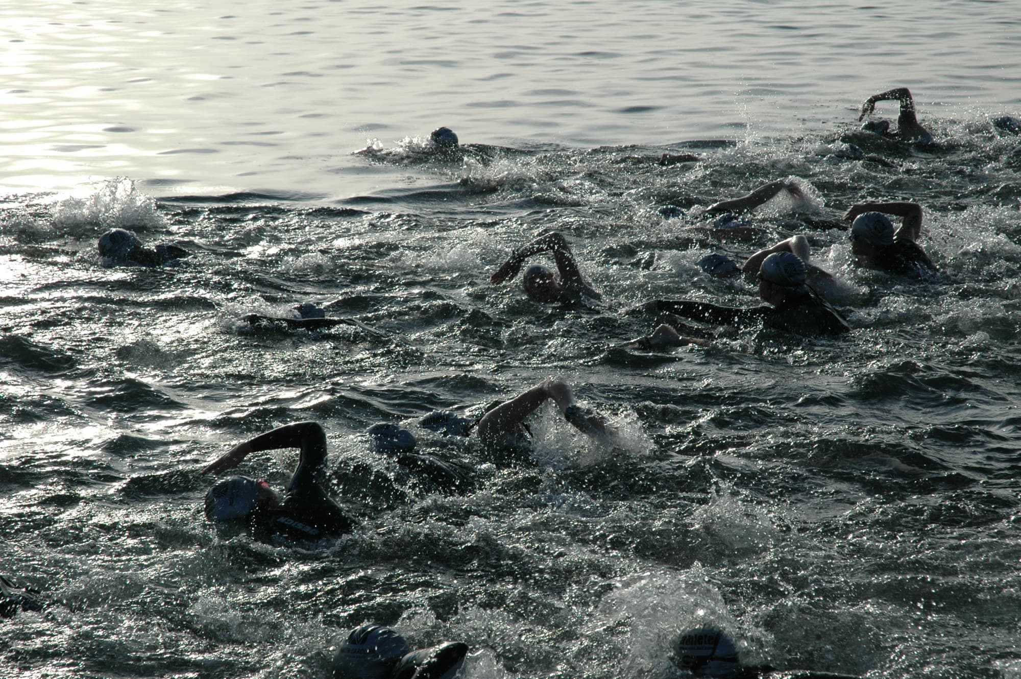 Dozens of swimmers in wetsuits and caps thrash through choppy grey water at the start of a triathlon, arms swinging over in every direction, with the low sun glinting off the lake behind them.