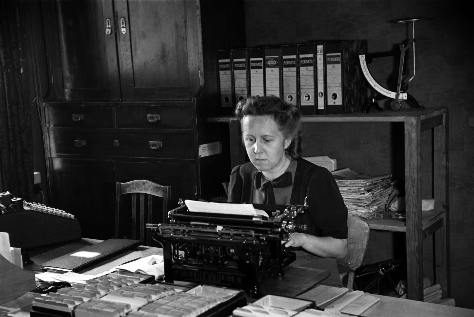 A black and white photograph from 1944 of a woman typing at a heavy desk typewriter in a printing house office. She sits on a hard chair with her shoulders rounded forward and her head reaching towards the page, shelves of files behind her.