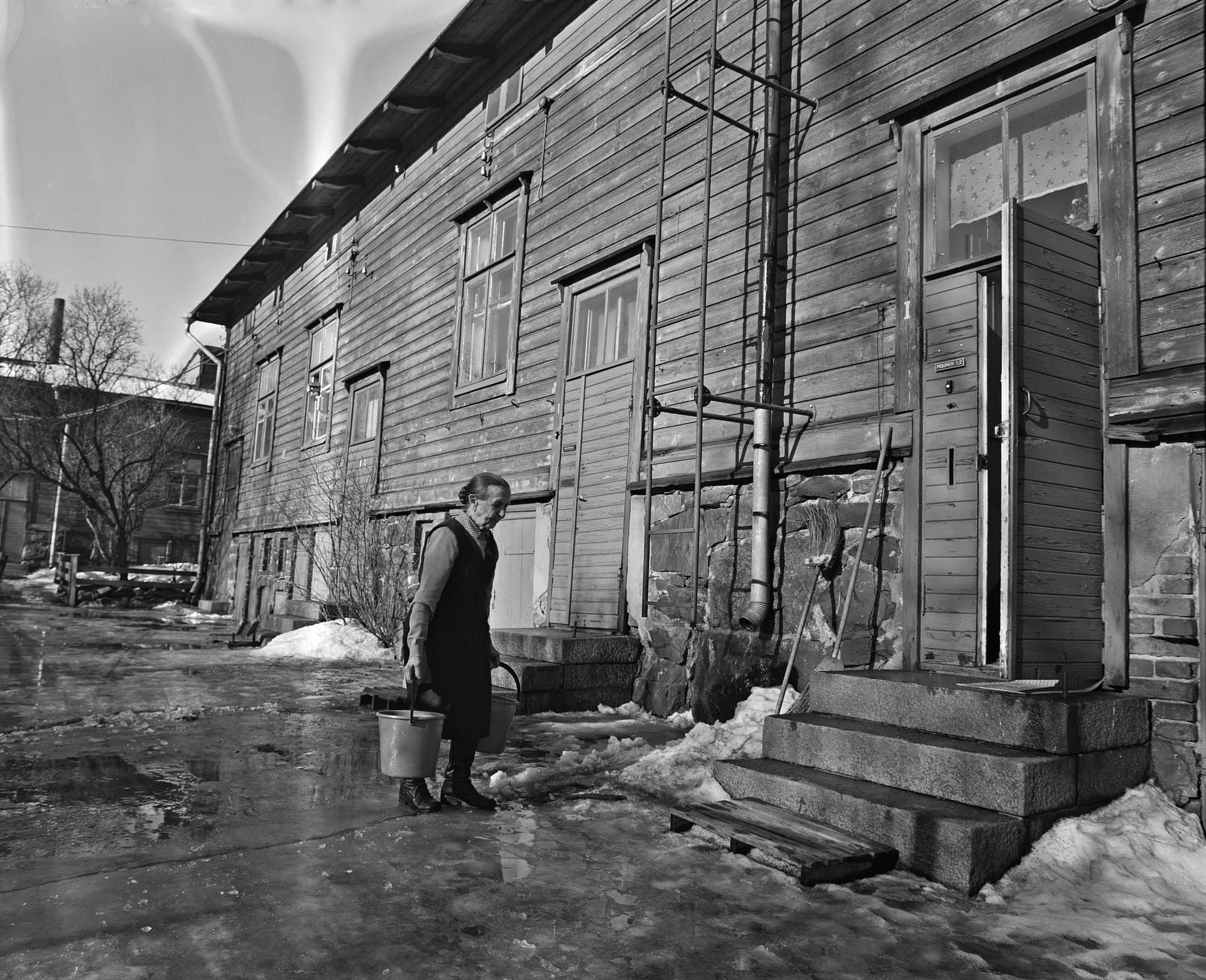 A black and white photograph from 1974 of a woman crossing a slushy yard beside a long wooden house, a full bucket of water hanging from each hand. She walks tall between the two weights, shoulders level, towards the steps of her door.