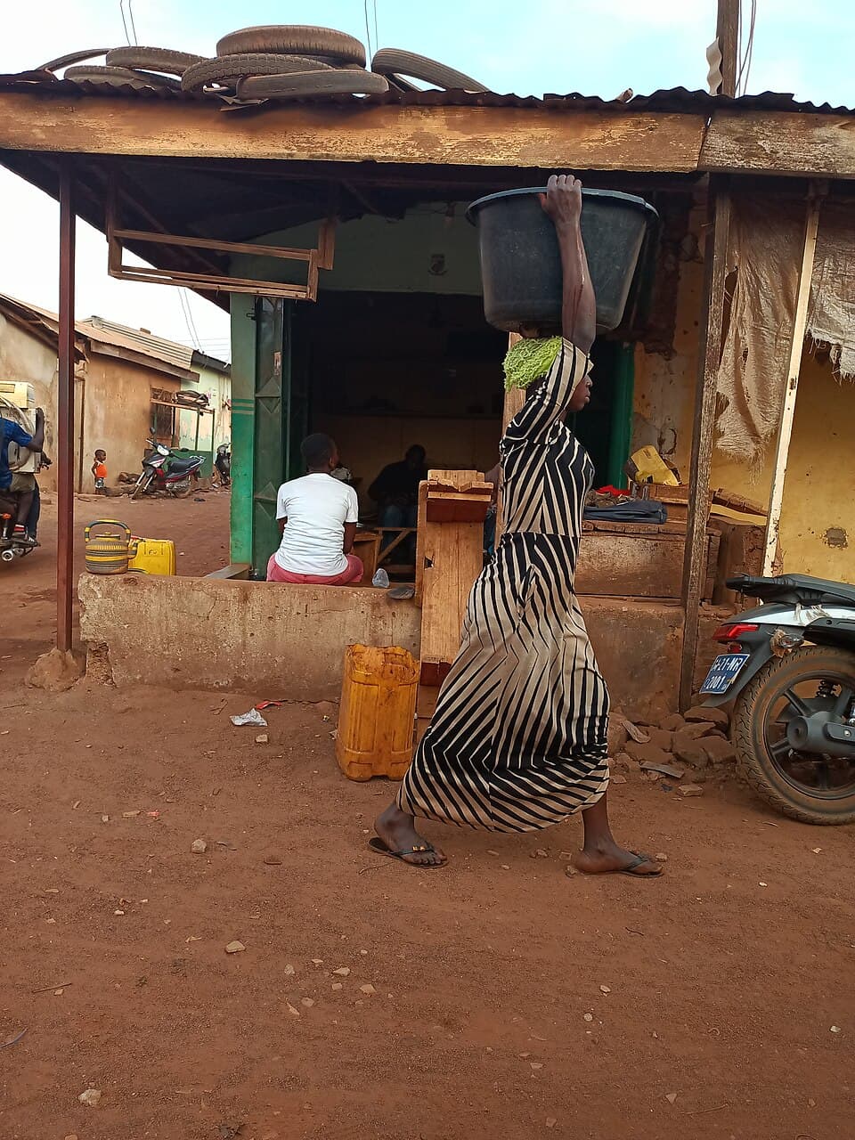A woman in a long striped dress walks in flip-flops along a dusty street in northern Ghana, seen from the side, steadying a large black tub of water on her head with one hand. Her ear sits over her shoulder, her shoulder over her hip, in one upright line.